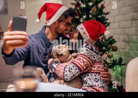 Coppia che prende auto ritratto con il loro cane di fronte all'albero di Natale. Sui cappelli di babbo natale. Concetto di vacanze di Natale. Foto Stock