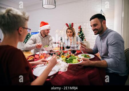 Due giovani coppie che cenano insieme a natale. Seduto al tavolo decorato pieno di cibo e divertirsi. Foto Stock