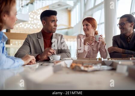 Un gruppo di dipendenti in una pausa gustando caffè e cibo in un'atmosfera allegra presso la mensa dell'azienda. Persone, lavoro, azienda, business concept. Foto Stock
