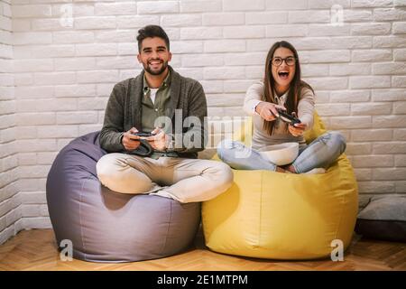 Coppia che gioca video gioco mentre si siede con gambe incrociate su borsa pigro. Joystick manuale. Concetto di vacanza invernale. Foto Stock