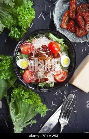 condimento di insalata di verdure con foglie di lattuga verde, pomodori ciliegini freschi e secchi, parmigiano grattugiato, uova, carne e crostini, in un piatto nero Foto Stock