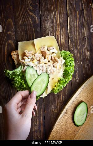 flatlay con le verdure a mano su sandwich con foglie di lattuga verde, formaggio, tonno a fette, cetrioli e pane di grano, al buio di legno Foto Stock