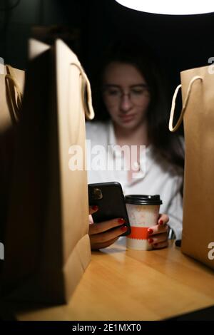 una giovane donna si siede tra borse di carta con acquisti e guarda il telefono. Foto Stock