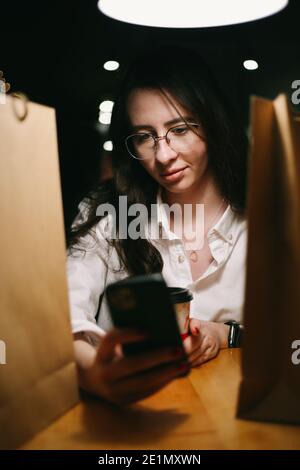 una giovane donna si siede tra borse di carta con acquisti e. guarda il telefono Foto Stock