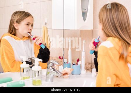 Tween ragazza che indossa pigiama arancione pennelli i suoi denti con elettrico spazzolino da denti in bagno Foto Stock