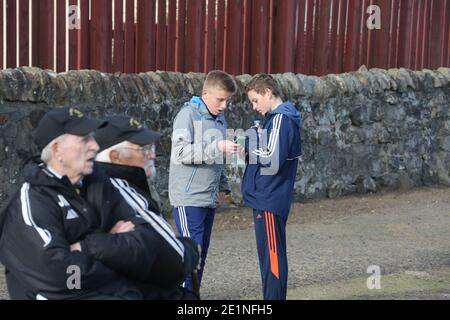 Due giovani ragazzi, ragazzi in una partita di calcio locale a Irvine, Ayrshire, Scozia, Regno Unito. Controllare i loro telefoni cellulari in primo piano due anziani supportati in matching treack tute Foto Stock