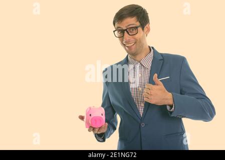 Studio shot di giovani happy businessman sorridere mentre dando pollice in alto e tenendo premuto salvadanaio Foto Stock