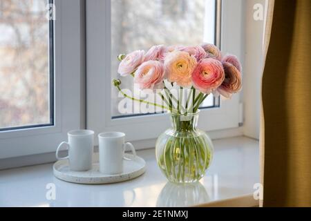 Bouquet di bicchierini Persiani rosa è in vaso rotondo di vetro su una soglia di finestra di pietra artificiale bianca dietro una tenda. Sul sono presenti due tazze bianche con tè Foto Stock