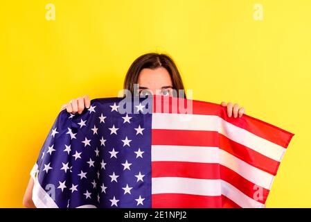 la giovane donna latina spaventa si copre con una bandiera americana nella paura delle deportazioni, isolata su sfondo giallo. Foto Stock
