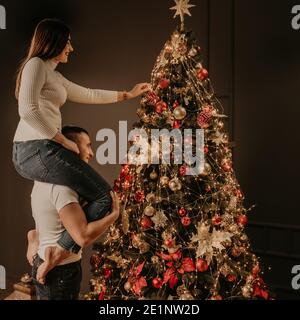 Giovane donna si siede sulle spalle posteriori dell'uomo e decora un albero di Natale, appende ornamenti. Casa decorata per Capodanno. Natale mattina. appartamento Foto Stock