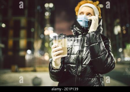 La donna adulta indossa una maschera di protezione e guanti medici mentre cammina lungo una città europea durante la quarantena, beve bevande e usa una cella Foto Stock