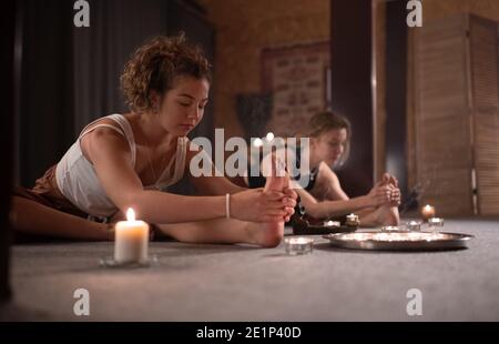 Allenatore femminile facendo Pascimottanasana posa variazione vicino a donna e candele Foto Stock