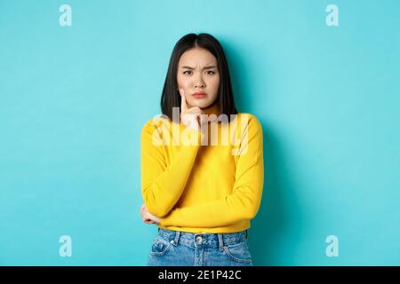 Immagine di una donna asiatica penosa e seria che tocca il mento, si accigliava e fissava la telecamera perplessa, rendendo difficile la scelta, in piedi su sfondo blu Foto Stock