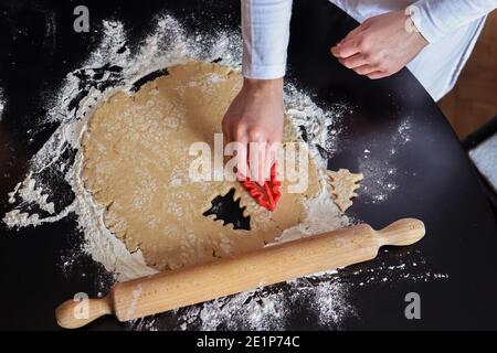 Mano della ragazza con stampo rosso del biscotto che taglia l'impasto nella forma di un albero di Natale su una scrivania con un perno di rotolamento e la farina. Preparazione e decorazione fatti in casa Foto Stock