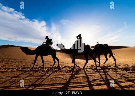 (Fuoco selettivo) vista mozzafiato della silhouette di due persone che cavalcano cammelli sulle dune di sabbia a Merzouga, Marocco. Foto Stock