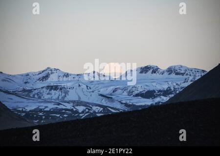 Luna piena che si erge su cime innevate di montagna ghiacciata vicino Solheimasandur DC3 relitto aereo roccia nera sabbia vulcanica Islanda del Sud Europa Foto Stock