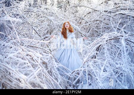 Giovane donna rossa, principessa, cammina in una foresta invernale in un vestito blu. Foto Stock