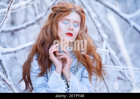 Giovane donna rossa, principessa, cammina in una foresta invernale in un vestito blu. Foto Stock