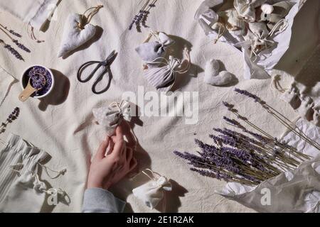 Sacchetti di lavanda fatti a mano in sacchetti di tessuto, cuori morbidi. Le mani fanno il sacchetto. Fiori secchi di lavanda per ripieno, forbici, candela. Piatto, tessuto Foto Stock