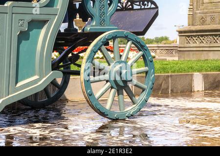 carrozza in legno verde con ruote in legno su pietre bagnate, sullo sfondo sono presenti aiuole e basi in pietra con aranciato intagliato Foto Stock