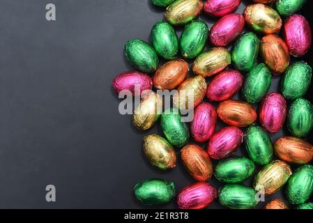 Uova pasquali multicolore avvolte in foglio di colore rosa, verde, arancione e giallo in un palo o in un gruppo, su sfondo grigio nero. Foto Stock