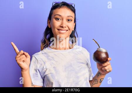 Giovane donna che beve l'infusione di compagno sorridente felice puntando con la mano e il dito a lato Foto Stock