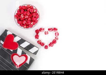 Concetto di film di San Valentino. Clapperboard con cuori, popcorn rosso caramello con spazio di copia su sfondo bianco. Foto Stock