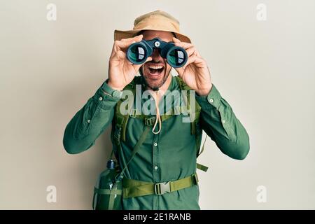 Bell'uomo con la barba che indossa il cappello explorer guardando attraverso binocoli sorridente e ridendo forte perché divertente scherzo pazzo. Foto Stock