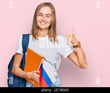 Bella giovane donna bionda scambio studente tenendo la bandiera russa sorridendo felice e positivo, pollice su facendo eccellente e segno di approvazione Foto Stock