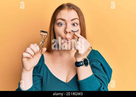 Giovane bella donna rossa che tiene arricciatore di ciglia e ciglia finte facendo faccia di pesce con la bocca e occhi squinting, pazzo e comico. Foto Stock