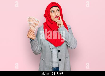 Bella donna ispanica che indossa hijab islamico tenendo in mano le banconote turche di lira faccia seria pensare alla domanda con la mano sul mento, pensieroso circa Foto Stock