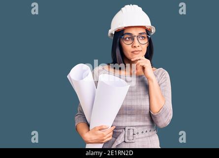 Giovane bella donna latina che indossa hardhat architetto e occhiali che tengono blueprints faccia seria pensare alla domanda con la mano sul mento, pensefu Foto Stock