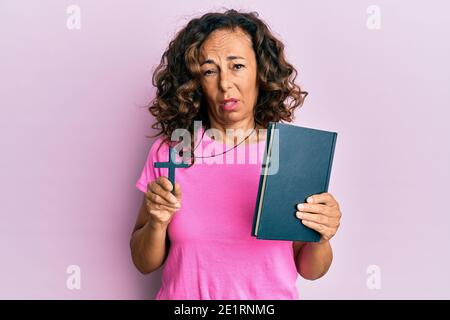 Donna ispanica di mezza età che tiene la bibbia e croce cristiana indistinta e confusa espressione. Concetto di dubbio. Foto Stock