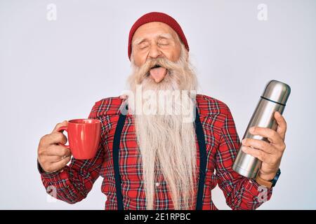 Vecchio uomo anziano con capelli grigi e barba lunga che tiene una tazza di caffè e la linguetta adesiva termica fuori felice con l'espressione divertente. Foto Stock