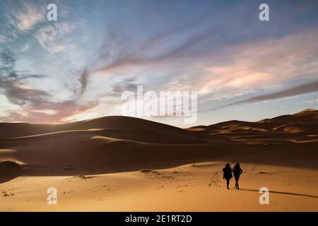 (Fuoco selettivo) Silhouette di due persone che camminano sulle dune di sabbia del deserto di Merzouga durante un tramonto mozzafiato. Merzouga, Marocco. Foto Stock