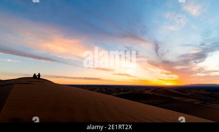 (Fuoco selettivo) Silhouette di una coppia seduta sulle dune di sabbia del deserto di Merzouga durante un tramonto mozzafiato. Merzouga, Marocco. Foto Stock