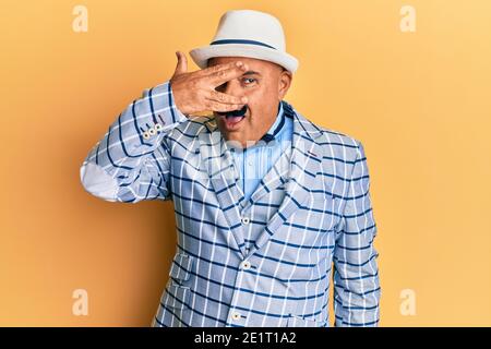 Uomo medio-orientale maturo con baffi che indossano vintage ed elegante stile di moda sbucciando in shock coprendo viso e occhi con la mano, guardando attraverso il fing Foto Stock