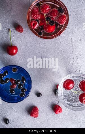 Bevanda fresca di acqua frizzante fredda con ciliegia, lampone e bacche di curry in tre vetri colorati su fondo di pietra in cemento, bevanda estiva di dieta Foto Stock