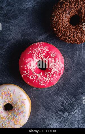 Ciambelle di cioccolato, rosa e vaniglia con spruzzi in fila, dessert con cibi dolci smaltati su fondo testurizzato in cemento scuro, vista dall'alto Foto Stock