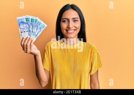 Giovane donna bruna che tiene in Sud africa 100 rand banconote cercando positivo e felice in piedi e sorridente con un sorriso fiducioso mostrare i denti Foto Stock