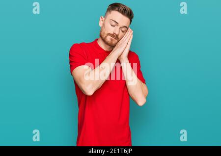 Giovane uomo rosso che indossa abiti casual dormendo stanco sognare e posare con le mani insieme mentre sorride con gli occhi chiusi. Foto Stock