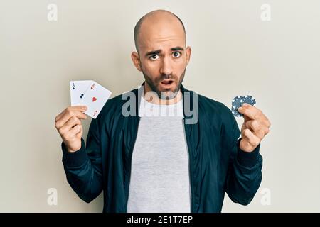 Giovane uomo ispanico che gioca a poker tenendo carte e chip da casinò in faccia shock, guardando scettico e sarcastico, sorpreso con bocca aperta Foto Stock