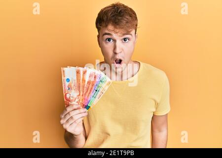 Giovane uomo caucasico che tiene le banconote di peso filippino spaventato e stupito con bocca aperta per sorpresa, faccia di incredulità Foto Stock