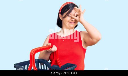 Donna bruna con sindrome di Down tenendo supermercato carrello di shopping sorridente buon fare ok segno con mano su occhio guardando attraverso dita Foto Stock