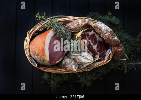 Cesto con salame, coppa e prosciutto su fondo di legno scuro. Specialità italiane. Decorazioni natalizie. Foto Stock