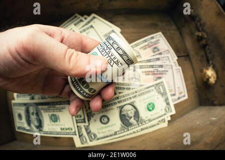 Mano che tiene cento banconote americane in rotolo sopra lo sfondo di cassetto di legno con lotto di dollari. Contanti di cento fatture del dollaro, valuta di soldi della carta. Foto Stock