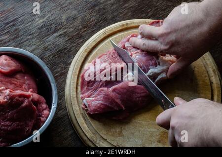 Cucina, concetto di ricette di carne. Le mani maschili tritare con un coltello grandi pezzi di manzo fresco crudo su un tagliere. Messa a fuoco selettiva. Foto Stock