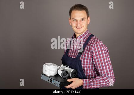 Ritratto di giovane elettricista allegro che tiene in mano il kit telecamera di sorveglianza parte anteriore di sfondo grigio Foto Stock