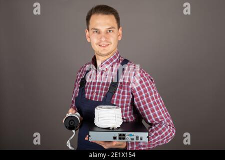 Ritratto di giovane elettricista allegro che tiene in mano il kit telecamera di sorveglianza parte anteriore di sfondo grigio Foto Stock