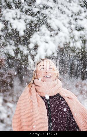 Donna bionda con cappotto viola e sciarpa rosa gode di nevicare e sfrigolarsi durante l'inverno in fondo alla foresta Foto Stock